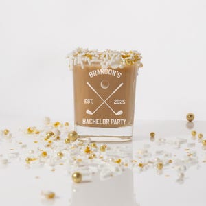 May include: A shot glass filled with a light brown liquid, rimmed with white sprinkles and gold sprinkles. The glass has the text "Brandon's EST. 2025 BACHELOR PARTY" with a golf-themed graphic. Gold and white sprinkles are scattered around the glass.