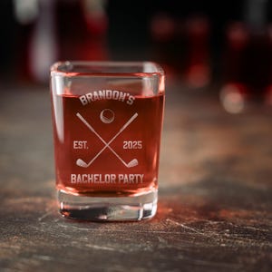 May include: A square shot glass filled with a red liquid. The glass is etched with the text "Brandon's" above a golf-themed design with crossed golf clubs and a golf ball. The text "EST. 2025" and "BACHELOR PARTY" are also etched on the glass.