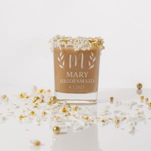 Pu&ograve; includere: Un bicchierino di vetro trasparente con un liquido marrone al suo interno. Il bicchiere ha una glassa bianca intorno al bordo ed &egrave; inciso con la lettera "M", il nome "Mary", la parola "Bridesmaid" e la data "4.3.2023".