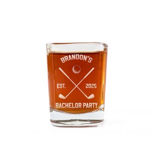 May include: A square shot glass filled with a brown liquid. The glass has white text and graphics, including the words "Brandon's Bachelor Party" and crossed golf clubs. The year "2025" is also displayed.