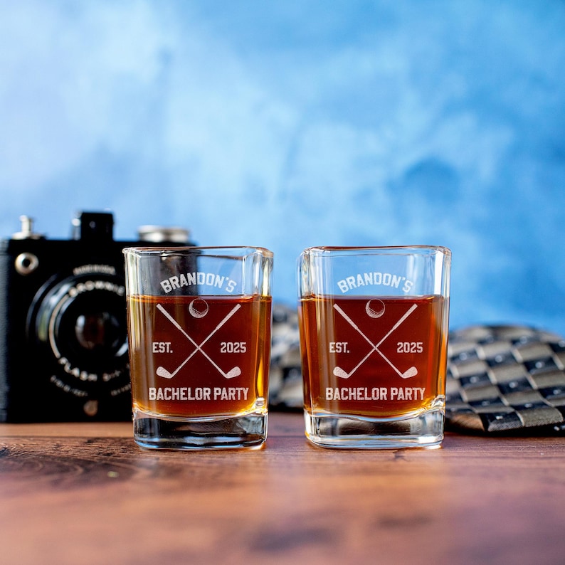 May include: Two square shot glasses filled with amber liquid. Each glass is etched with "Brandon's", a golf ball, crossed golf clubs, "EST. 2025", and "Bachelor Party". A vintage camera and patterned tie are in the background.