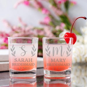 Pu&ograve; includere: Due bicchieri da shot in vetro trasparente con un liquido rosa al loro interno. I bicchieri sono incisi con le lettere "S" e "M" con un motivo floreale e il testo "Sarah Bridesmaid 4.3.2023" e "Mary Bridesmaid 4.3.2023".