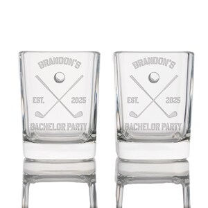 May include: Two clear shot glasses with etched golf-themed designs. Each glass features the text "Brandon's", a golf ball, crossed golf clubs, "EST. 2025", and "BACHELOR PARTY". The glasses are rectangular with a thick base.