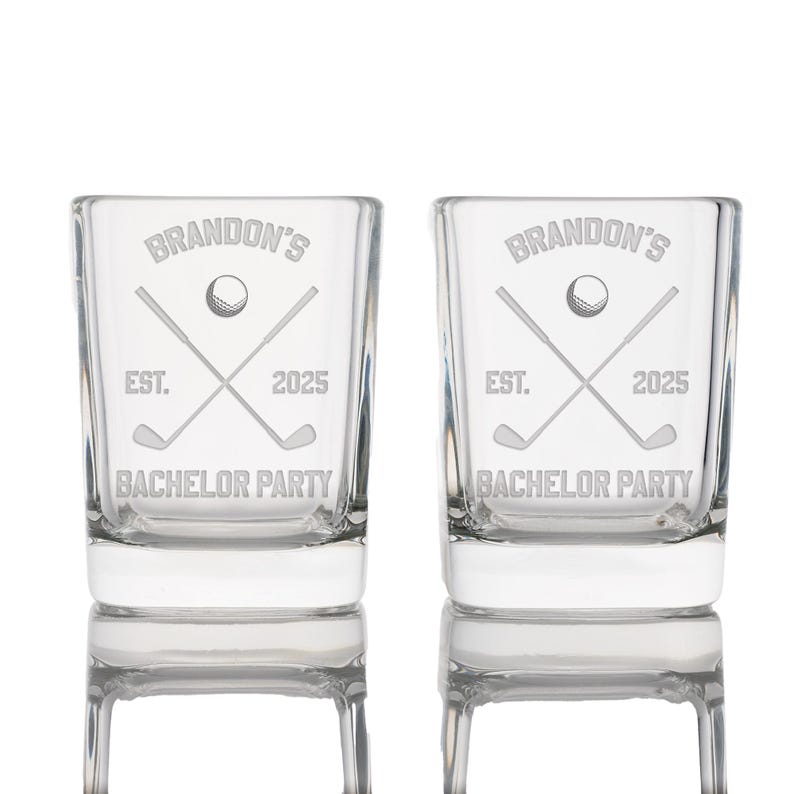 May include: Two clear shot glasses with etched golf-themed designs. Each glass features the text "Brandon's", a golf ball, crossed golf clubs, "EST. 2025", and "BACHELOR PARTY". The glasses are rectangular with a thick base.