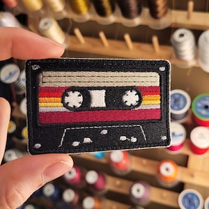 May include: Embroidered patch in the shape of a retro cassette tape. The patch features a black border, with a design of red, orange, and yellow stripes. The tape reels are white. A fun accessory for jackets, bags, or other fabric items.