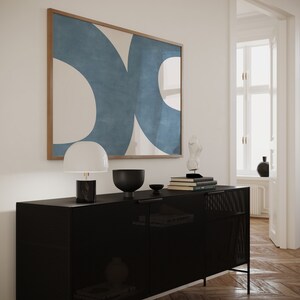 May include: A black metal cabinet with a mesh front and two doors. The cabinet is decorated with a white lamp, a black bowl, and a stack of books. A framed abstract painting with blue and white shapes hangs on the wall behind the cabinet.