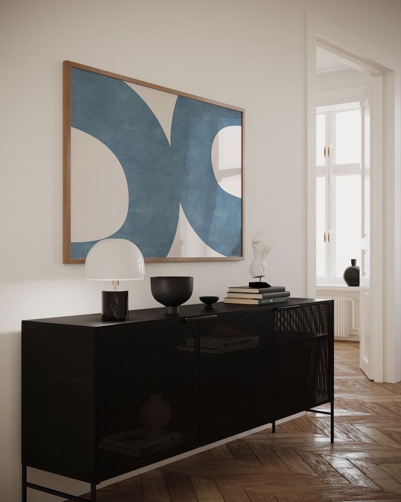 May include: A black metal cabinet with a mesh front and two doors. The cabinet is decorated with a white lamp, a black bowl, and a stack of books. A framed abstract painting with blue and white shapes hangs on the wall behind the cabinet.