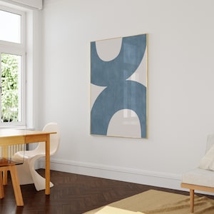May include: A minimalist abstract art print with a blue and white design in a gold frame. The print is hanging on a white wall in a room with a wooden floor.
