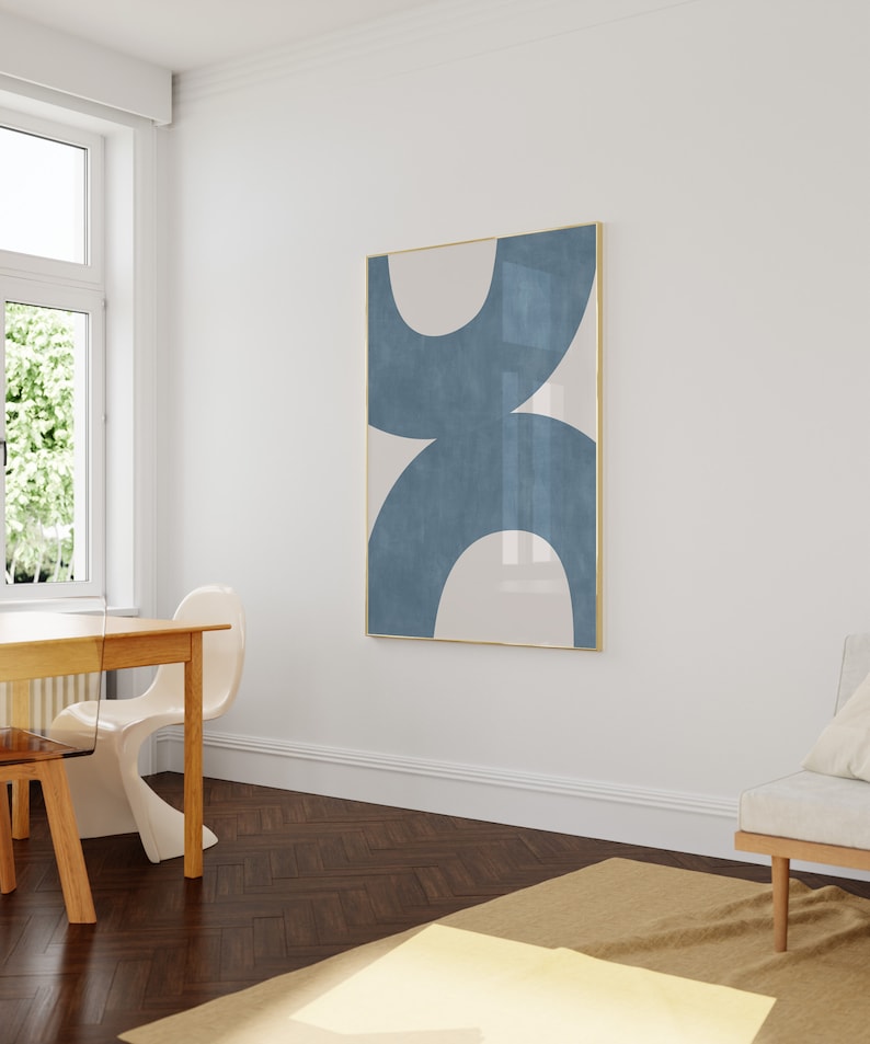 May include: A minimalist abstract art print with a blue and white design in a gold frame. The print is hanging on a white wall in a room with a wooden floor.