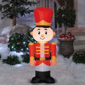 May include: An inflatable Christmas decoration shaped like a toy soldier. The soldier wears a red jacket with gold accents, a black belt, and a tall red hat. The soldier's face is white with a smiling expression. The decoration is standing in a snowy outdoor setting.