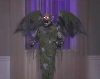 May include: A Halloween decoration of a monstrous figure with large wings. The figure is green and grey, with tattered clothing and glowing red eyes. The creature's mouth is open in a scream, creating a scary effect.