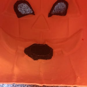 May include: Close-up of an orange plastic jack-o'-lantern mask. The mask has black mesh eye and mouth openings. The face is carved with a triangular nose and a smiling mouth. The mask is a Halloween decoration.
