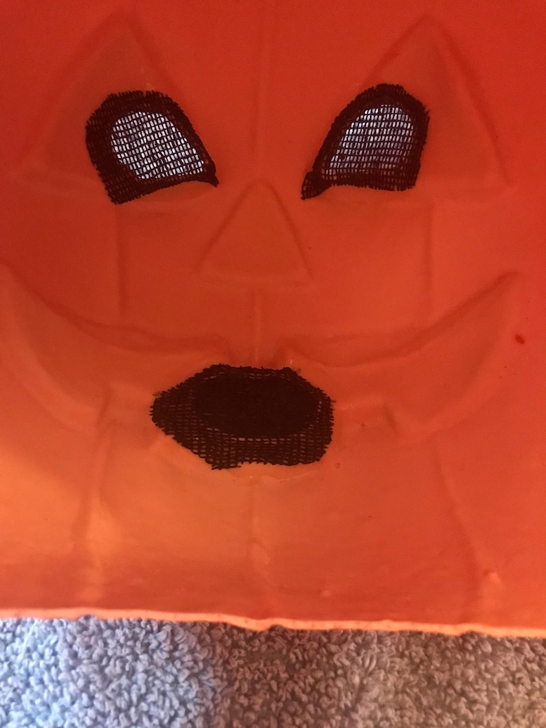 May include: Close-up of an orange plastic jack-o'-lantern mask. The mask has black mesh eye and mouth openings. The face is carved with a triangular nose and a smiling mouth. The mask is a Halloween decoration.