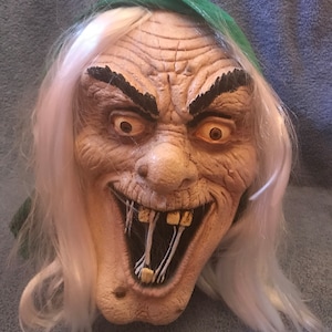 May include: A grotesque Halloween mask with a wrinkled face, exaggerated features, and a sinister grin. The mask has white hair, dark eyebrows, and a green hood. The teeth are yellowed and the eyes are wide and staring.