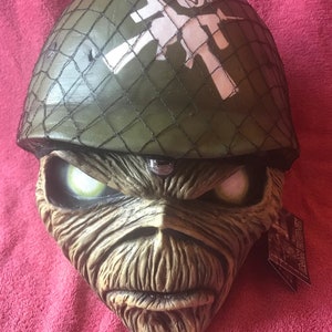 May include: A green army helmet with a white skull and crossbones design. The helmet is worn by a green and brown face mask with glowing green eyes. The mask is a replica of the face of Eddie, the mascot of the heavy metal band Iron Maiden.