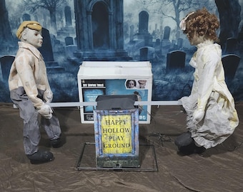 May include: A Halloween animated seesaw with two doll figures in vintage clothing. The dolls are positioned on either side of a seesaw. The base of the seesaw has the text "HAPPY HOLLOW PLAY GROUND". The background depicts a graveyard scene.