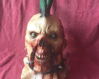 May include: A grotesque Halloween mask with a punk-style green mohawk. The mask features a decaying face with exposed teeth, blood, and green eyes. A studded collar adds to the menacing appearance. The mask is designed for costume use.