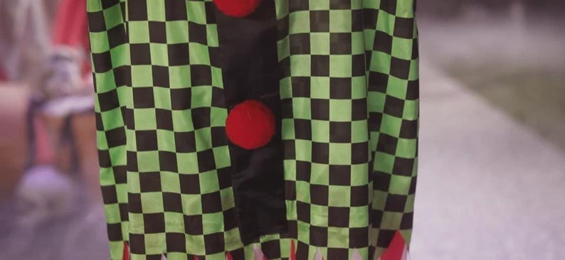 May include: A costume featuring a green and black checkered pattern. The costume has three large red pom-pom buttons down the front. The bottom of the costume has red fringe.