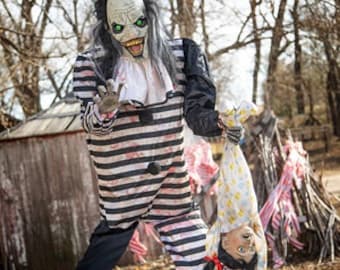 May include: A Halloween decoration depicting a sinister clown with a pale face, green eyes, and a wide, grinning mouth. The clown is dressed in a black and white striped costume and holds a child upside down. The backdrop features trees and a rustic wooden building.