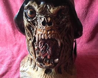 May include: A grotesque, detailed, brown and black latex mask of a zombie wearing a black German military helmet. The mask has a wide open mouth with exposed teeth and a red tongue.