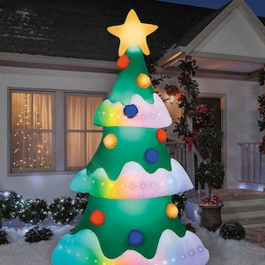 May include: An inflatable Christmas tree with a glowing yellow star on top. The green tree has white snow-like accents and colorful lights. It is decorated with orange, yellow, and blue ornaments. The tree is set up outside a house at night.