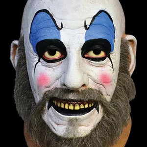May include: A white and blue clown mask with a gray beard and a wide, toothy grin. The mask has blue painted eyebrows and red cheeks.