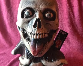 May include: A white skull mask with a large, red tongue sticking out. The mask has a realistic look with detailed bone structure and blue eyes. The mask is on a stand with a bone-like design.