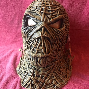 May include: A brown and tan mask with a woven texture and two eye holes. The mask has a textured, detailed design that resembles a face.