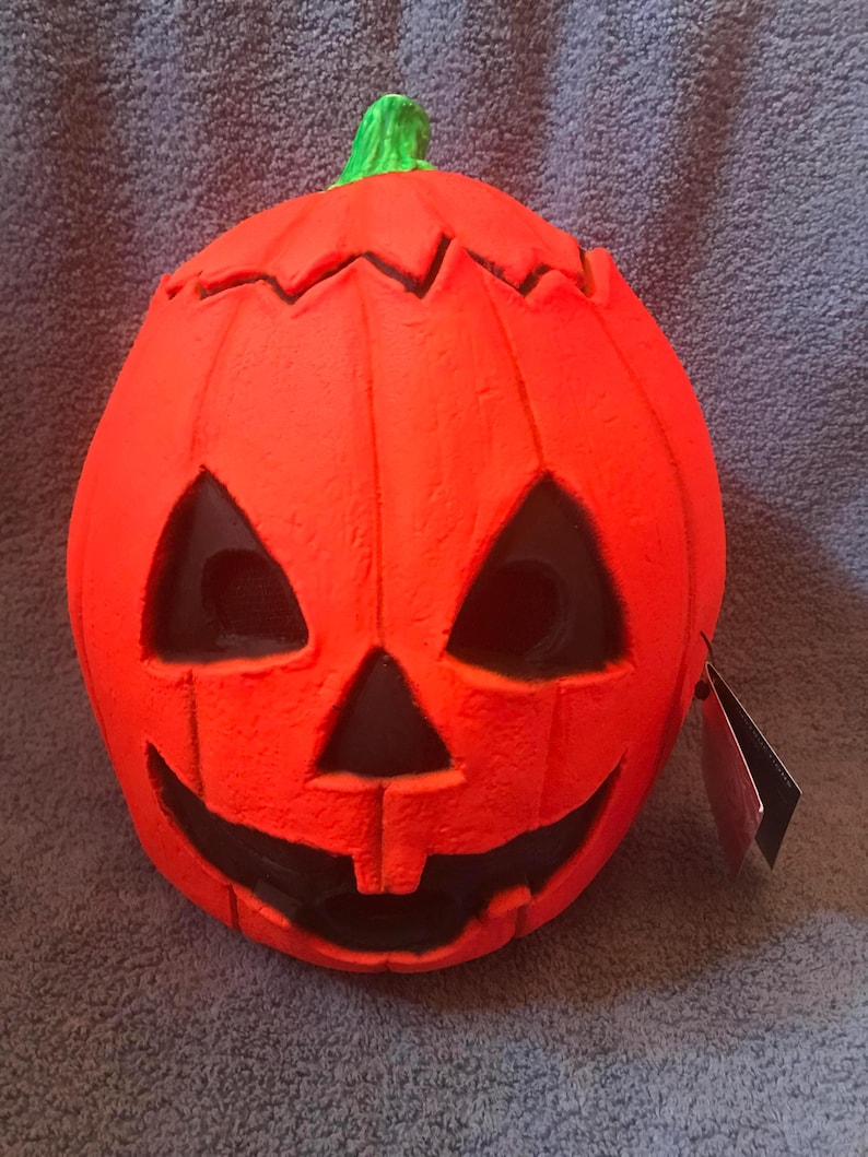 May include: A bright orange Halloween pumpkin mask with a carved face featuring triangular eyes and a wide, toothy grin. The pumpkin has a green stem and a textured surface. A tag is attached to the side.