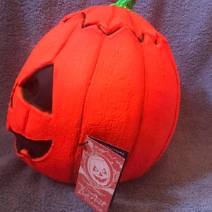 May include: A bright orange Halloween pumpkin with a carved face and a green stem. The pumpkin has a tag that reads "Don Post Studios" and features a pumpkin illustration. The pumpkin is a festive decoration.