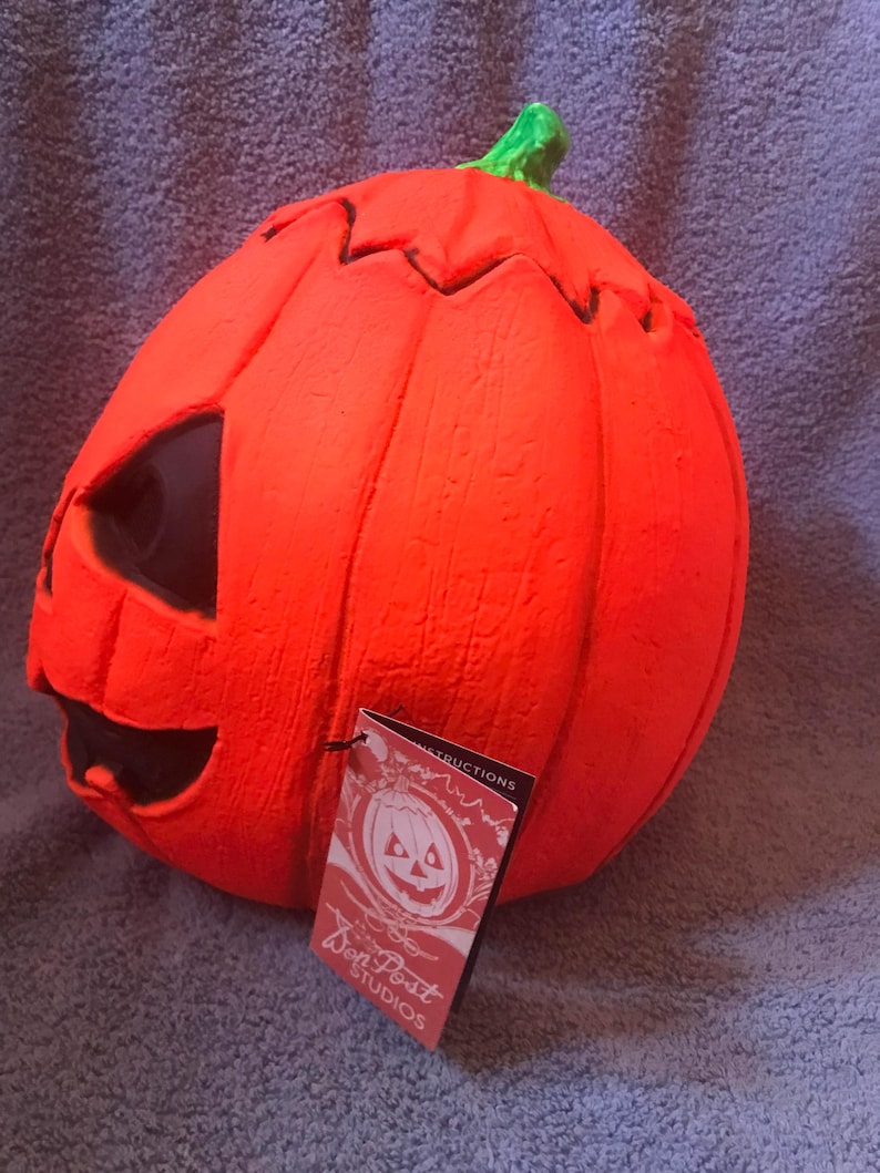May include: A bright orange Halloween pumpkin with a carved face and a green stem. The pumpkin has a tag that reads "Don Post Studios" and features a pumpkin illustration. The pumpkin is a festive decoration.