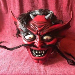 May include: A red devil mask with horns, a black mustache, and a wide, toothy grin. The mask has a black and red painted design and large, white eyes.