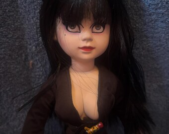 May include: A doll with black hair and a black dress. The doll has a fair complexion, red lipstick, and dramatic eye makeup. The dress has a plunging neckline and a belt with a decorative buckle. The doll's hair is styled in a bouffant.
