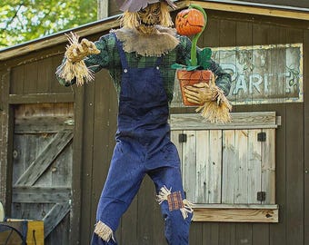May include: A Halloween scarecrow decoration stands on green grass, wearing a plaid shirt, blue overalls, and a straw hat. It holds a potted pumpkin. The scarecrow has a burlap collar and straw hands. Patches are sewn on the knees of the overalls.