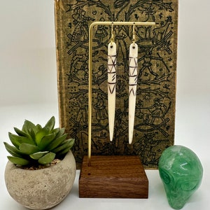 May include: A pair of long, white bone earrings with a geometric pattern etched in black. The earrings are hanging from a gold stand against a vintage book background.