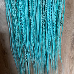 May include: A collection of turquoise dreadlocks, of varying thicknesses and textures, suspended from a wooden hanger. Some dreads are smooth, others are braided or twisted, creating a visually interesting display. The colour is a vibrant, solid turquoise.