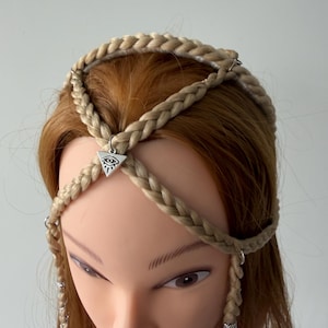 May include: A blonde braided hair accessory featuring a silver-toned triangular charm with an eye symbol at the centre. The accessory consists of multiple interconnected braids that form a crown-like structure, accented with small silver rings.