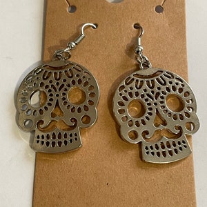 May include: A pair of silver sugar skull earrings with intricate cutouts. The earrings are on a brown card.