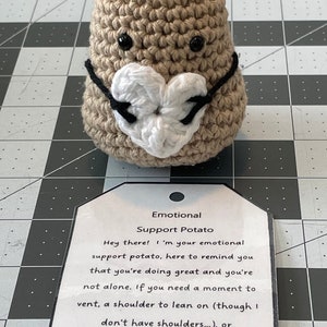 May include: A crocheted potato wearing a heart-shaped mask with the text "Emotional Support Potato" on a tag. The tag reads "Hey there! I'm your emotional support potato, here to remind you that you're doing great and you're not alone. If you need a moment to vent, a shoulder to lean on (though I don't have shoulders...), or just someone to listen. I'm here for you. You've got this!"