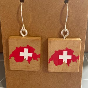 May include: A pair of wooden earrings with a red and white Swiss flag design. The earrings are square shaped and have silver hooks.