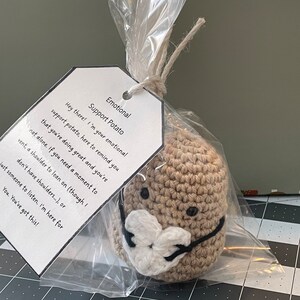 May include: A crocheted brown potato with a white crocheted face mask. The potato is packaged in a clear plastic bag with a tag that reads "Emotional Support Potato" and includes a message of encouragement.