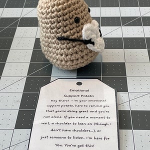 May include: A crocheted potato wearing a white face mask with the text "Emotional Support Potato" on a tag. The tag reads "Hey there! I'm your emotional support potato, here to remind you that you're doing great and you're not alone. If you need a moment to vent, a shoulder to lean on (though I don't have shoulders...), or just someone to listen. I'm here for You. You've got this!"