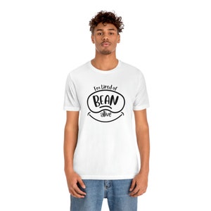 May include: White t-shirt with the text "I'm tired of BEAN alive" in black. The text is inside a black oval with a smiling mouth graphic below. The shirt is short-sleeved and worn by a person.