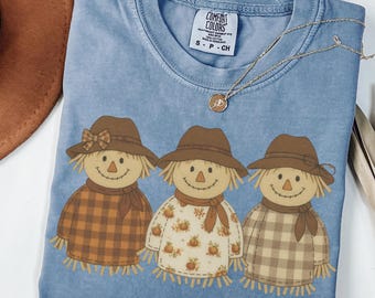 May include: A blue Comfort Colors t-shirt featuring a graphic of three scarecrows wearing hats and scarves. The scarecrows have straw hair and are wearing different patterned shirts. A gold necklace with a pendant is draped across the shirt.