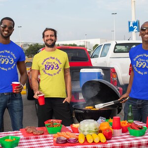 May include: Three friends wearing blue, yellow, and blue t-shirts with the text "Academy High School Class of 1993 Celebrating 30 years" in gold and purple. They are gathered around a table with a grill and food, celebrating their high school reunion.