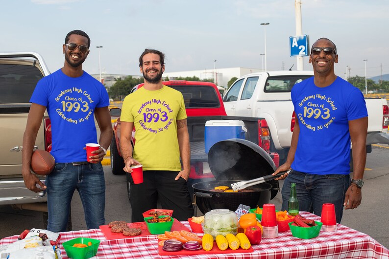 May include: Three friends wearing blue, yellow, and blue t-shirts with the text "Academy High School Class of 1993 Celebrating 30 years" in gold and purple. They are gathered around a table with a grill and food, celebrating their high school reunion.