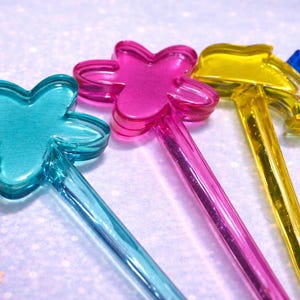 May include: Four colourful plastic wands are displayed in a close-up shot. The wands are transparent and feature different shapes: a flower, a star, and a hammer. The colours are teal, pink, yellow, and blue. The background is light purple with star confetti.