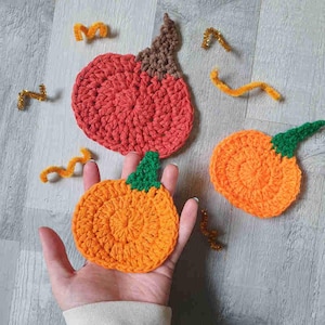Könnte beinhalten: Handgehäkelte Kürbisse in Orange, Rot und Braun mit grünen Stielen. Die Kürbisse sind auf einer hellen Holzmaserung mit goldenen und orangefarbenen Pfeifenreinigern angeordnet. Eine Hand hält einen der Kürbisse.