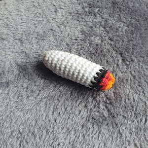 Peut inclure: Un jouet en forme de cigarette crocheté avec un corps blanc et un embout rouge, orange et noir.