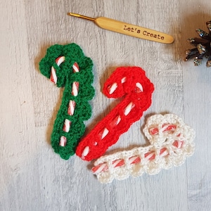 May include: Three crocheted candy cane ornaments in green, red and white. The ornaments are made with a white yarn and have a red and white striped candy cane design. The ornaments are on a wooden surface with a crochet hook and a pine cone.